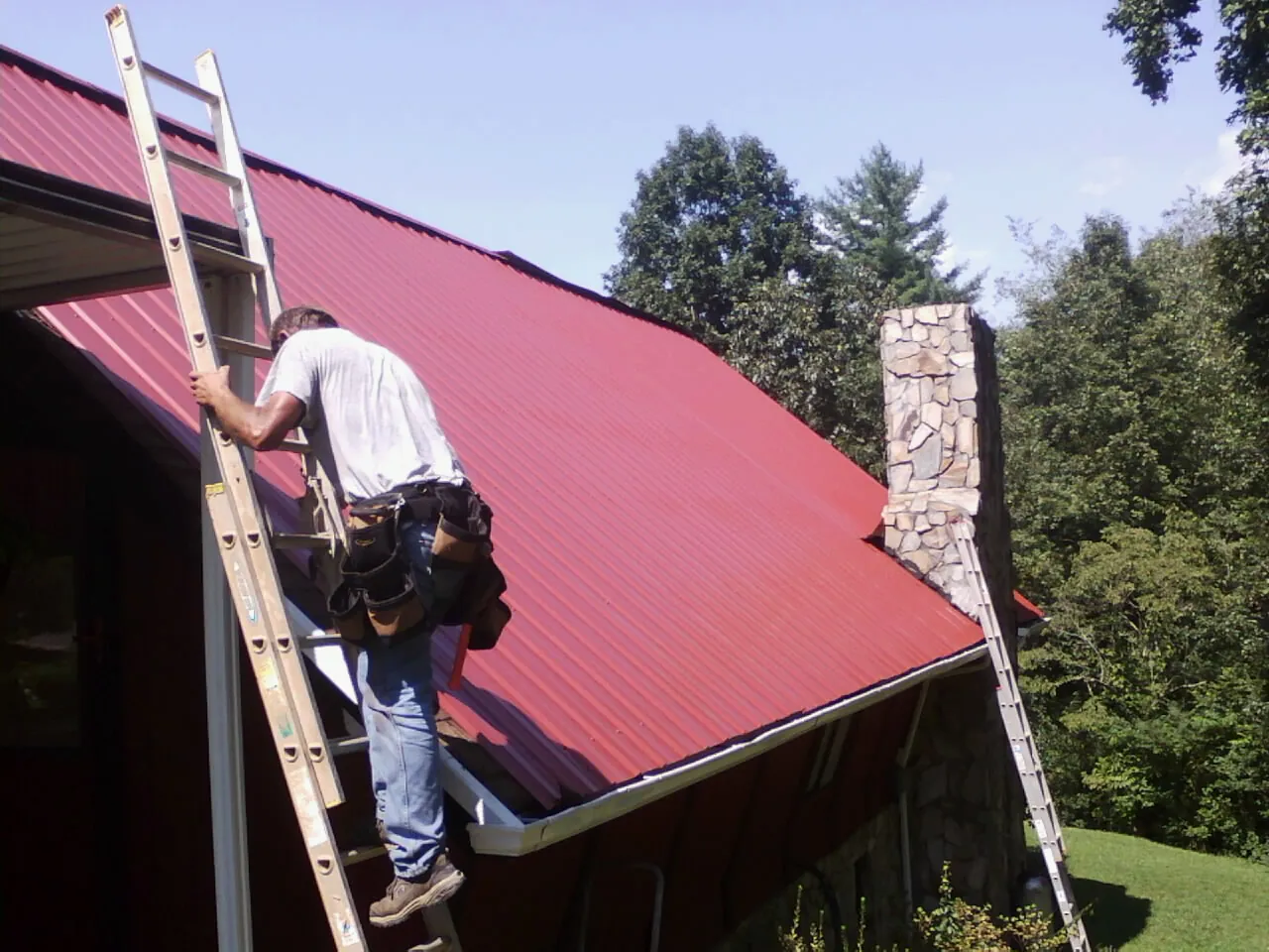 Roofer on a ladder beside a red metal roof and stone chimney — Soffit & Fascia Repair by Roofing Brothers in Hamlin